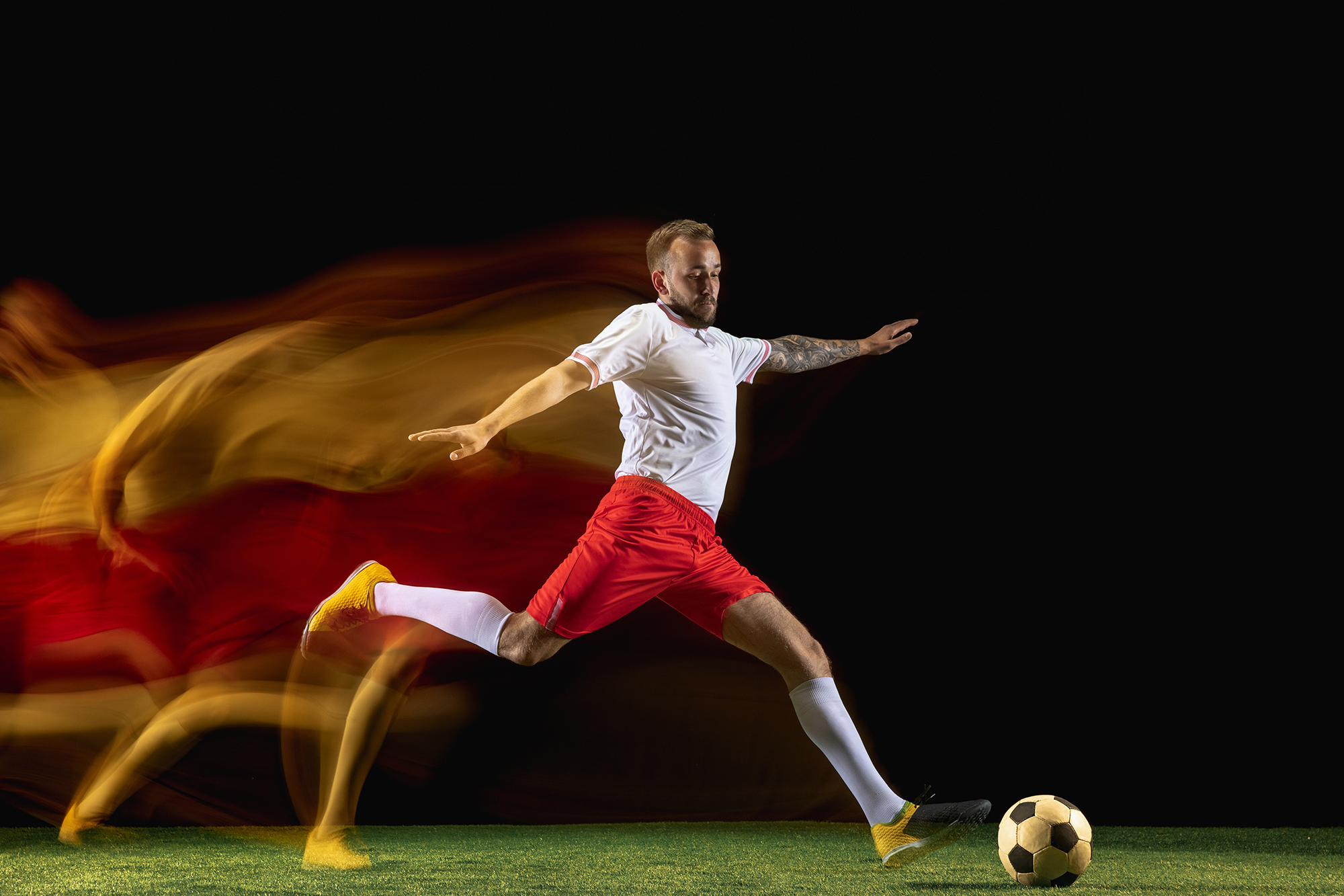 One step. Young caucasian male football or soccer player in sportwear and boots kicking ball for the goal in mixed light on dark background. Concept of healthy lifestyle, professional sport, hobby.