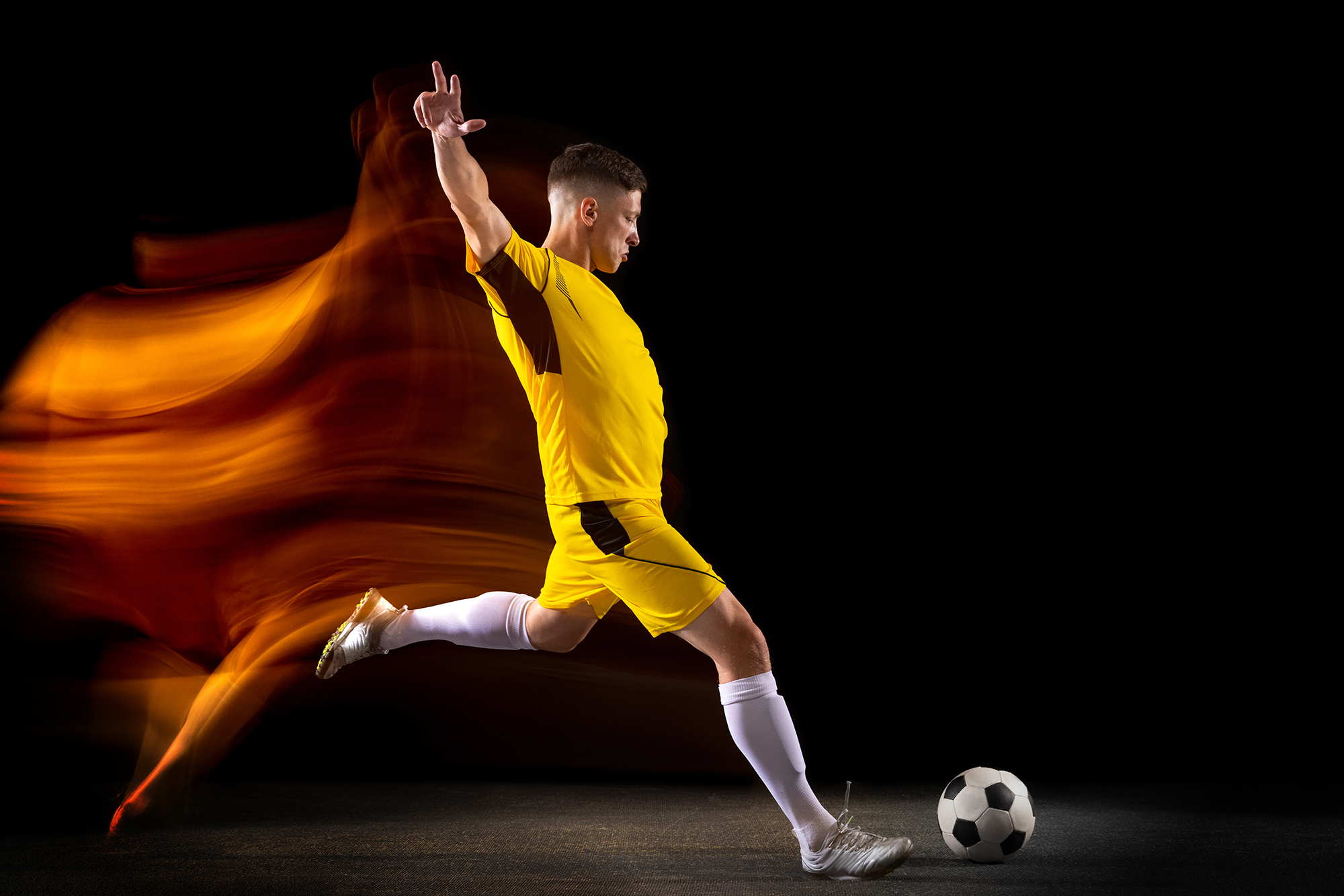 Champion. Young caucasian male football or soccer player in sportwear and boots kicking ball for the goal in mixed light on dark background. Concept of healthy lifestyle, professional sport, hobby.